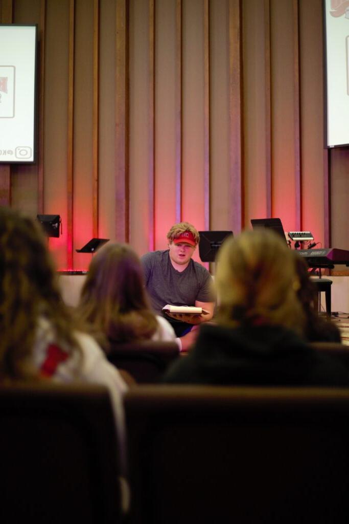 A person wearing a red visor speaks to an audience in a dimly lit room with red backlighting and vertical wooden panels.